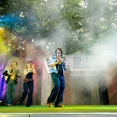 Theaterszenenfoto mit vier Schauspielern, die als Musikgruppe mit verschiedenen Instrumenten und Kostümen im Stil der 70er Jahre auftreten.
