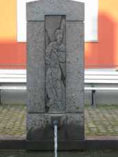 Brunnenstock aus Basalt mit einem Bronzerelief des Heiligen Andreas mit seinem typischen Attribut, dem Kreuz.